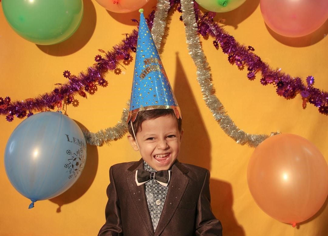 A young boy wearing a party hat and a colorful tux poses in front of some balloons and garland