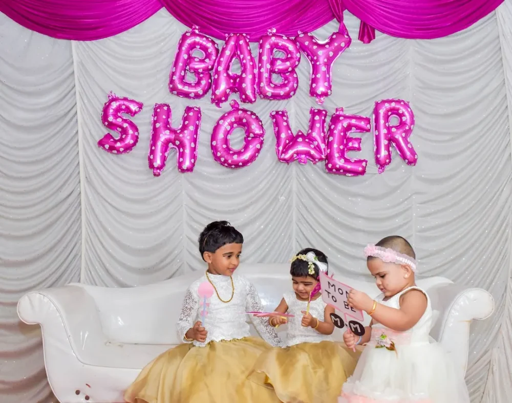 Three children pose against a backdrop featuring balloons that read "BABY SHOWER" at an event.