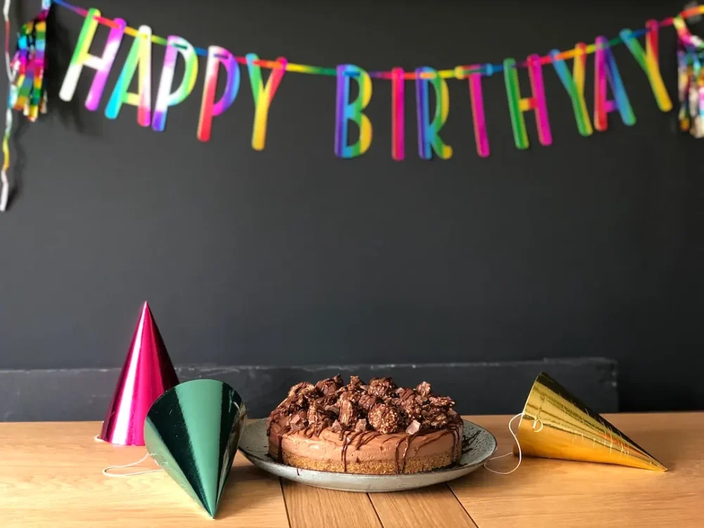 A birthday cake sits on a table with a few party hats and a "Happy Birthday" sign hanging above it.
