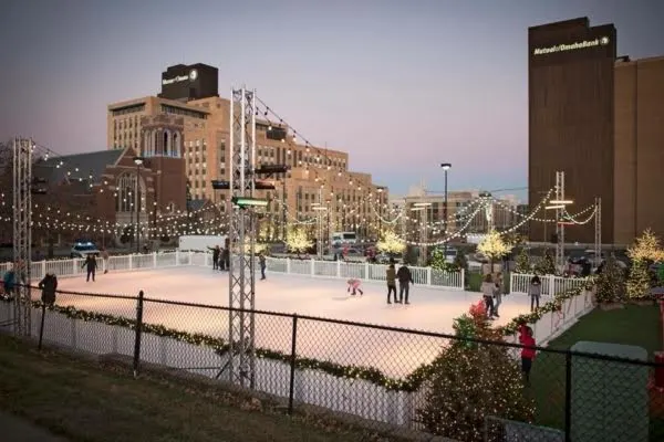 Large outdoor fenced ice skating rink in city downtown with skaters and clear draped rink lights.