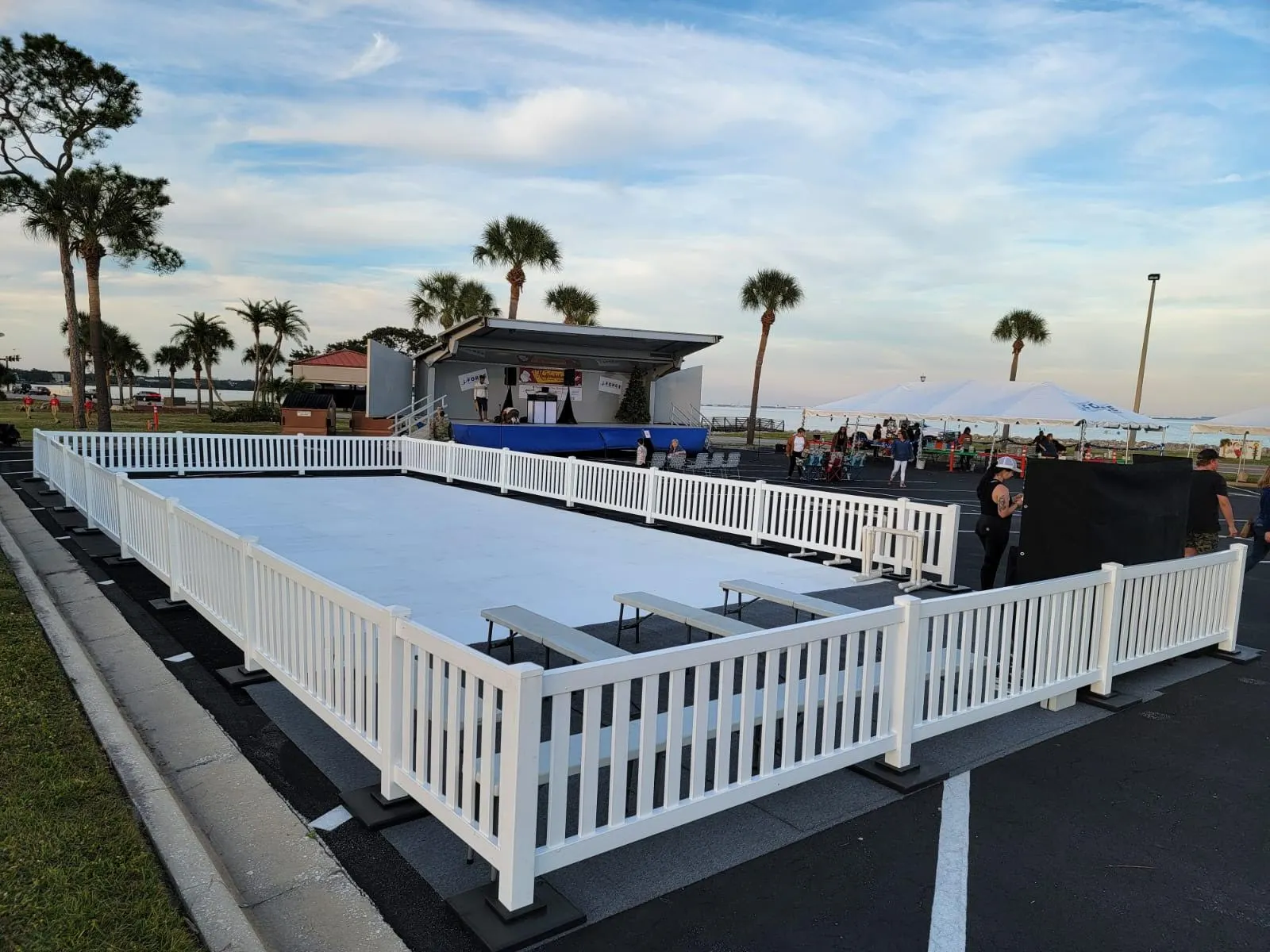Outdoor ice rink with white fence next to palm trees, festival stage and tents.