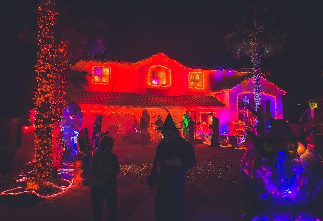 People in costumes mingle outside a house that is elaborately decorated for Halloween with lights, props, and other accessories.
