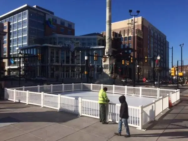 Small outdoor ice skating rink at downtown city intersection with white fence and two people.