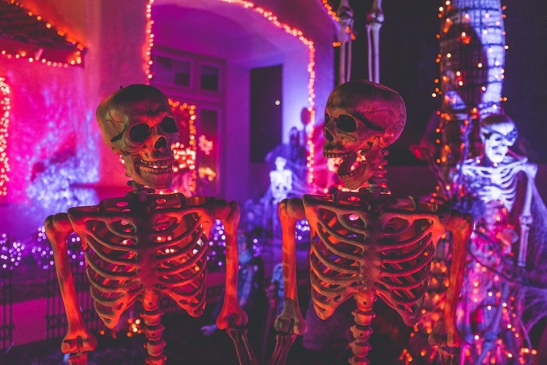 Two skeletons stand in front of a house that is elaborately lit and decorated for Halloween.